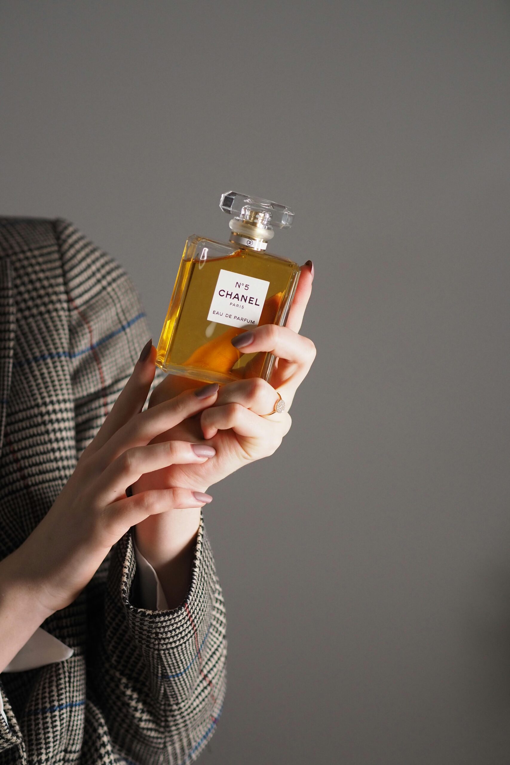 Close-up of hands holding Chanel No. 5 perfume bottle against a soft gray background.